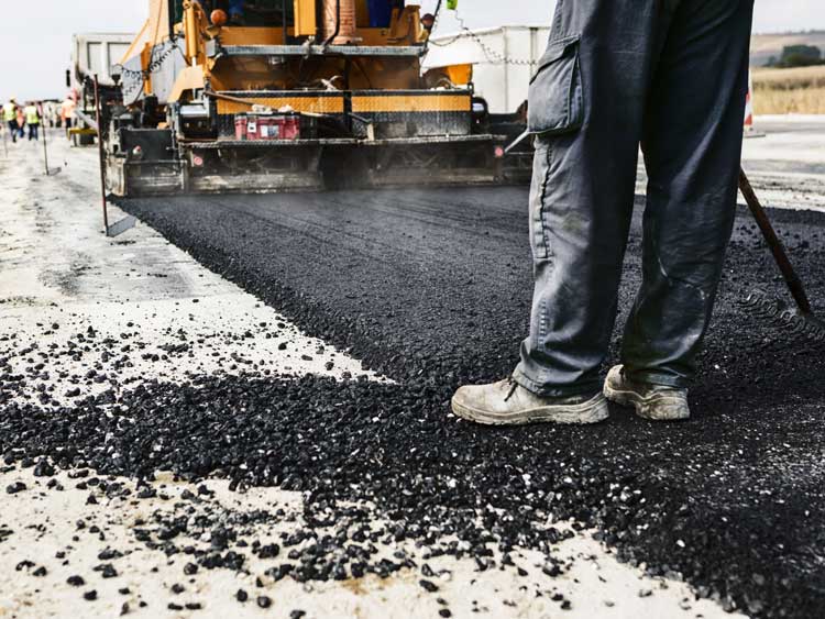 A road worker stands on a newly asphalted road as a road surfacing machine continues to pave the road.
